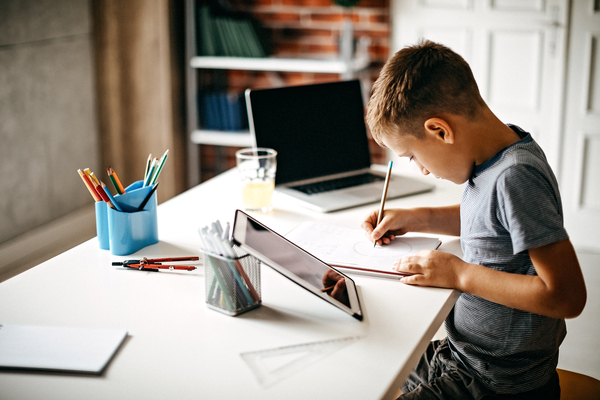 Un niño haciendo uso tarea en una mesa junto a una tableta y un ordenador portátil